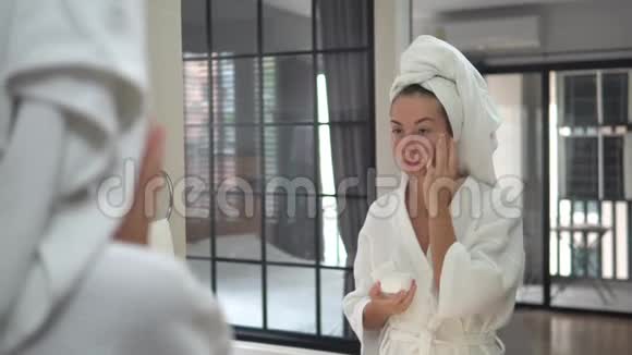 美丽的年轻女子头上戴着毛巾在镜子前集中在淋浴后的皮肤上给她的脸涂上奶油视频的预览图