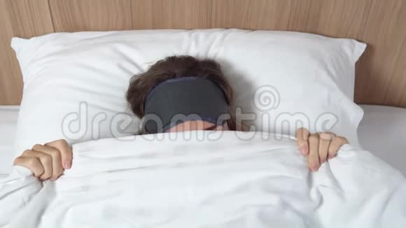 可爱的棕色眼睛的年轻女性躲在白色的毯子下头上戴着睡眠面具左看右看角度很高视频的预览图
