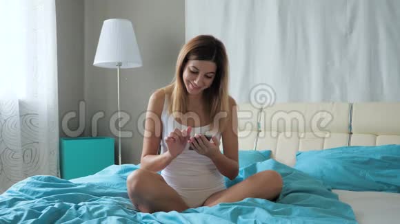 早上坐在床上的漂亮女人使用智能手机4k视频的预览图