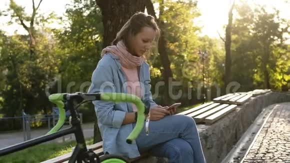 体贴的女孩坐在城市公园的长凳上旁边有她徒步旅行的自行车看着她的手机穿着视频的预览图