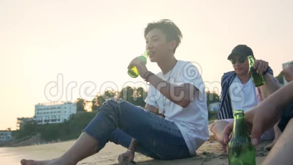 年轻的亚洲成年人坐在沙滩上放松喝啤酒视频的预览图