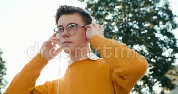 在阳光明媚的街道上戴着眼镜戴着耳机欣赏音乐的年轻可爱金发少年的肖像4k视频的预览图