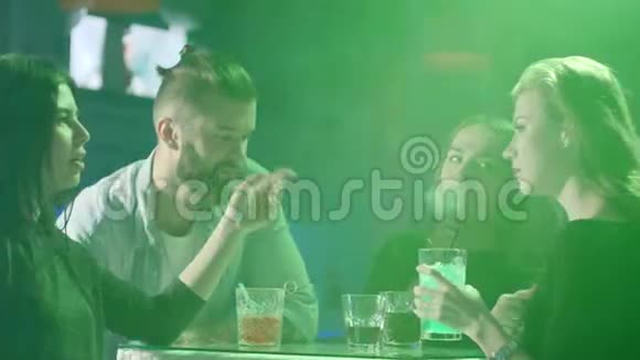 年轻的朋友在派对上的彩灯背景下在桌子旁玩着五颜六色的饮料视频的预览图