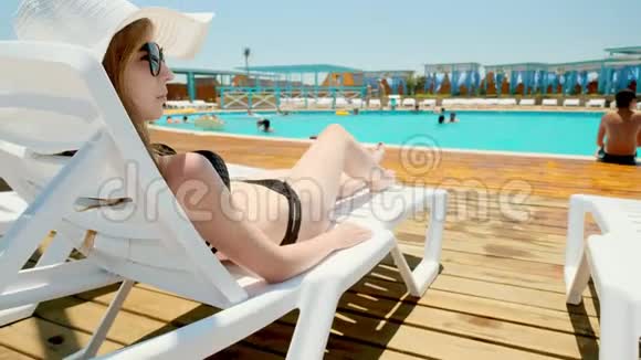 一个穿着黑色泳衣和一顶白帽子的快乐女孩躺在外面游泳池附近的休息室里他钉了一顶帽子真正的视频的预览图