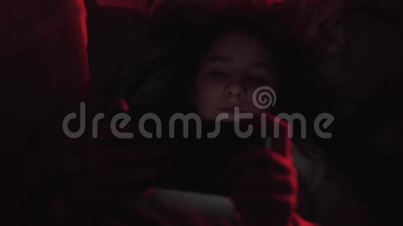 小女孩躺在黑暗的毯子下看着智能手机房间里有红灯视频的预览图