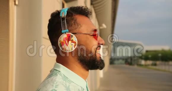 穿着时尚粉色太阳镜和耳机的放松的美国黑人男子正在机场航站楼附近大笑4k个镜头视频的预览图