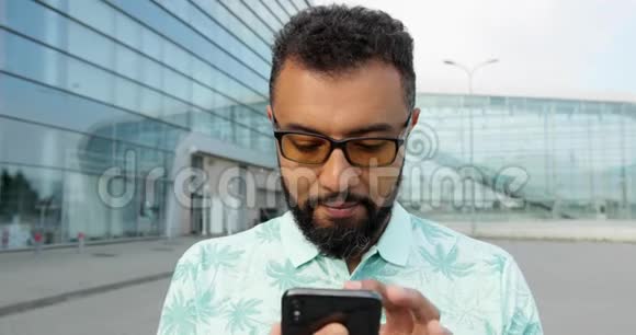 严肃而自信的美国黑人戴眼镜的男子正在玻璃大楼上使用手机他是视频的预览图