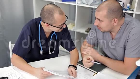 年轻的医生坐在办公室的桌子旁讨论治疗和研究笔记本上的笔记视频的预览图
