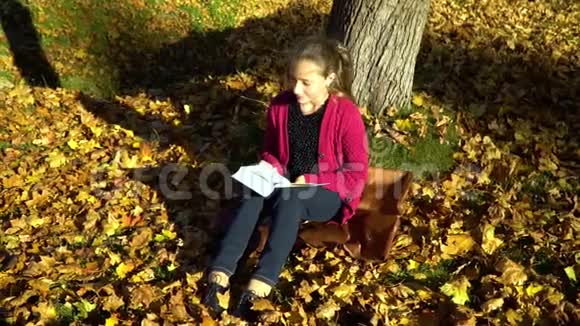 一个年轻漂亮的十几岁的女孩坐在秋天森林的一棵树下读一本书她做梦女孩很安详视频的预览图