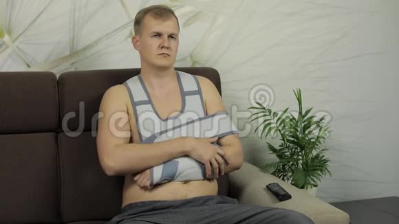 一个痛苦的男人胳膊断了戴着手臂支撑坐在沙发上看电视视频的预览图