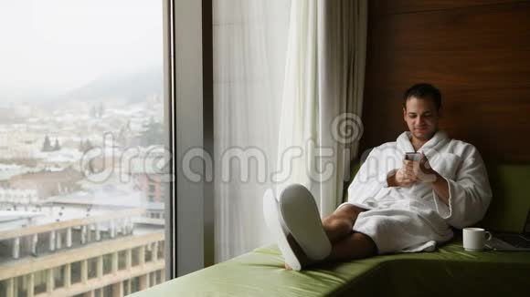 年轻的微笑英俊的男人穿着浴袍在手机上发短信微笑着他坐在靠近窗户的地方视频的预览图