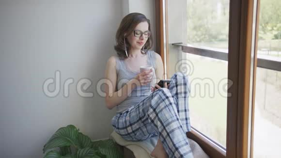 微笑的女人坐在窗户上用智能手机和耳机视频的预览图