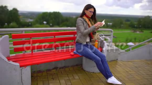 夏天带着眼镜的年轻美女坐在公园外面的长凳上看书4千克视频的预览图