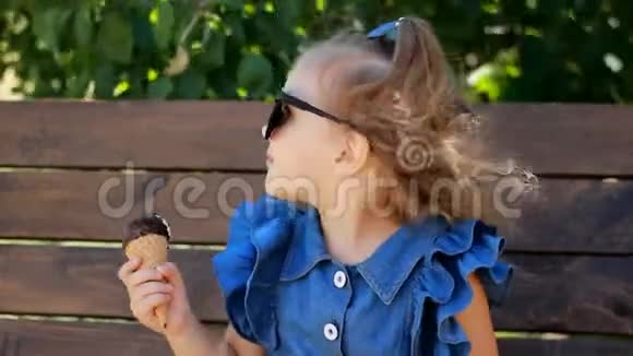 一个阳光明媚的夏日戴太阳镜的小女孩正坐在公园的长凳上吃冰淇淋一幅肖像画视频的预览图