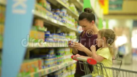 有着白种人外表的可爱女孩正在吃棒棒糖坐在超市的手推车里有背景的母亲选择视频的预览图