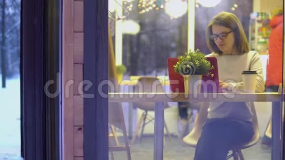 深思熟虑的概念年轻女孩穿着白色毛衣戴着眼镜在咖啡馆里用红色笔记本电脑工作查看视频的预览图