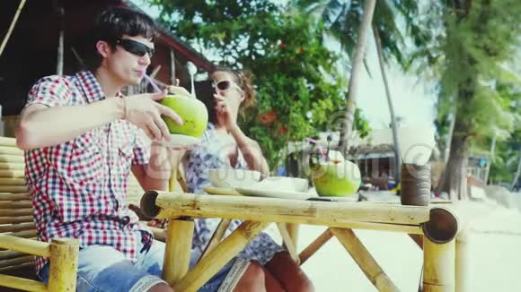 一对戴着太阳镜的年轻夫妇坐在海边的一家漂亮的咖啡馆里女孩用手机给男友拍照视频的预览图