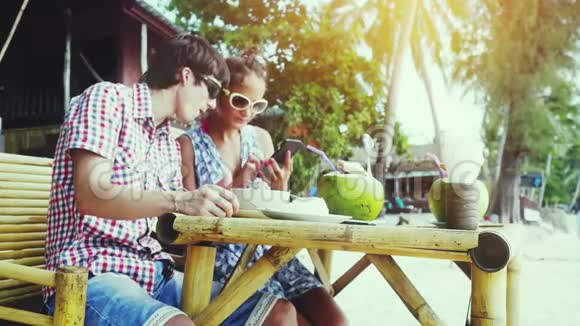 一对戴着太阳镜的年轻夫妇坐在海边的一家漂亮的咖啡馆里带男人看手机的女孩视频的预览图