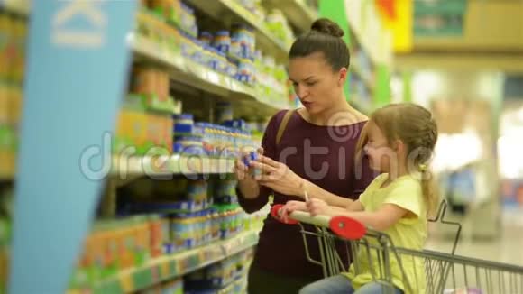 有着白种人外表的可爱女孩正在吃棒棒糖坐在超市的手推车里有背景的母亲选择视频的预览图