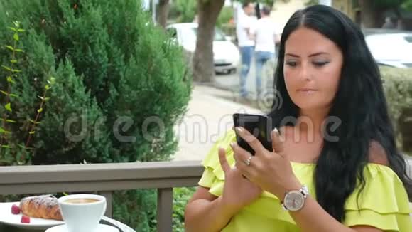 年轻漂亮的黑发女人坐在户外咖啡馆里喝咖啡用电话视频的预览图