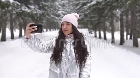 女孩站在冬天的森林里用智能手机自拍视频的预览图