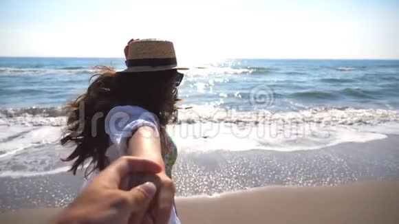 年轻的女人把男朋友拉到海边享受暑假或假期戴帽子的女孩握着男性的手视频的预览图