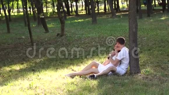 快乐的一对夫妇坐在公园的草地上拥抱着倚在树上视频的预览图