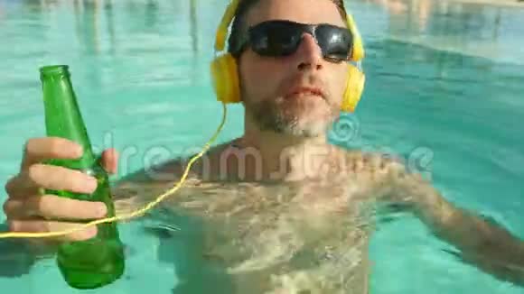 年轻快乐有魅力的男人带耳机听音乐在热带度假游泳池喝啤酒视频的预览图