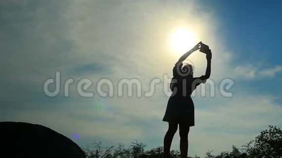 年轻女子穿着裙子站在岩石上用手机拍照的剪影慢动作1920x1080视频的预览图