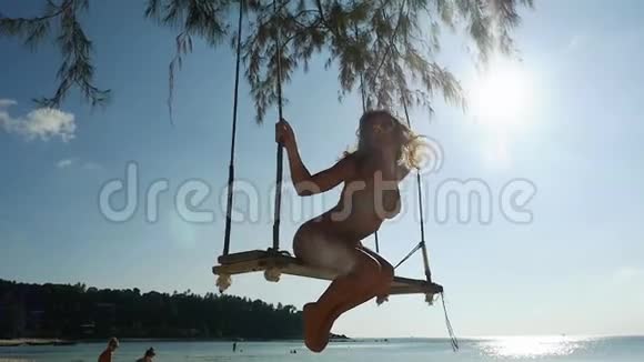 戴着太阳镜和比基尼的年轻迷人女子正在热带海滩的秋千上荡秋千身上放着闪亮的镜头视频的预览图