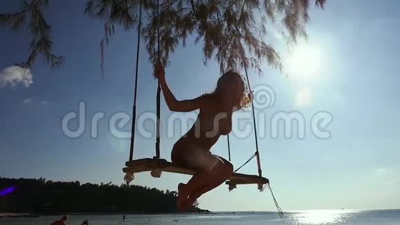 戴着太阳镜和比基尼的漂亮女孩在热带海滩的秋千上放松有着闪亮的镜头Lare效果慢慢慢慢视频的预览图