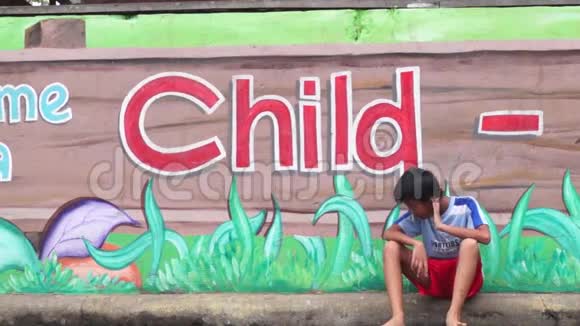 男孩坐在壁画混凝土学校墙的人行道上等待他的玩伴视频的预览图