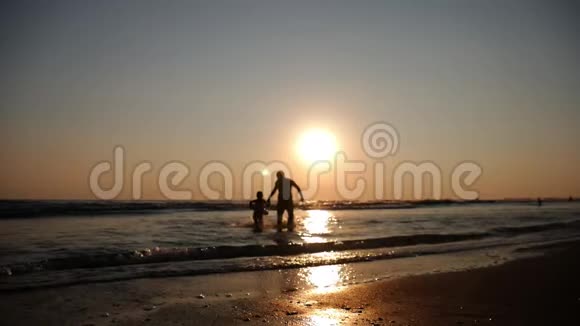 父亲和他的小女儿的剪影在日落时快乐地跑进大海溅到他们身上视频的预览图