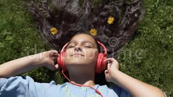 迷人的女孩留着长长的黑发躺在公园的草地上听着耳机上的音乐慢动作高清高清视频的预览图