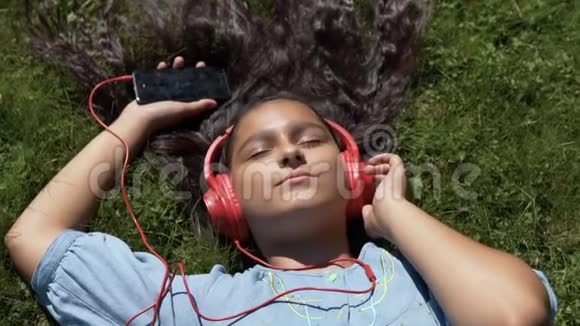 有魅力的长发女孩躺在公园的草地上用智能手机用红色耳机听音乐视频的预览图