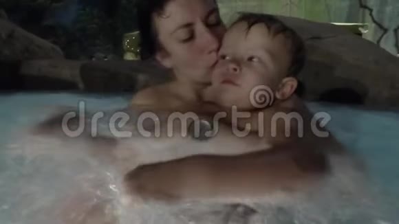 快乐的年轻妈妈抱着小儿子微笑着和他在游泳池里玩视频的预览图