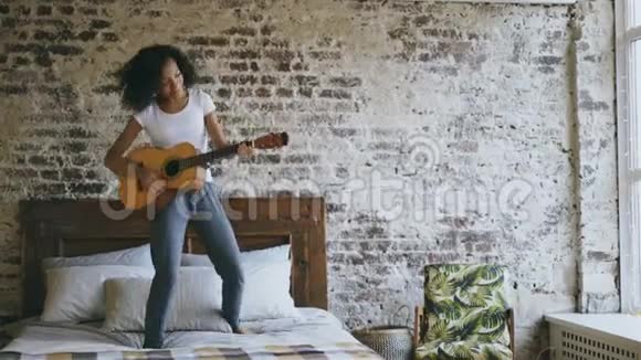 混合种族的年轻滑稽女孩唱歌和弹声吉他同时在家里的床上尽情跳舞视频的预览图