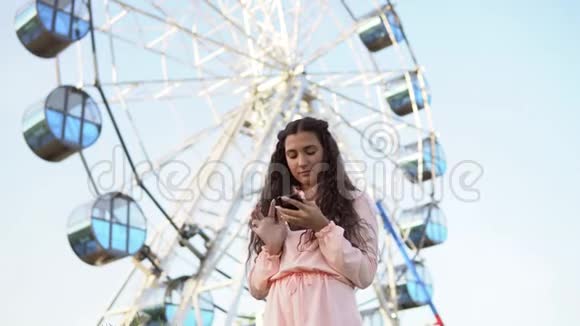这个女孩正在使用一部智能手机站在摩天轮附近4K视频的预览图