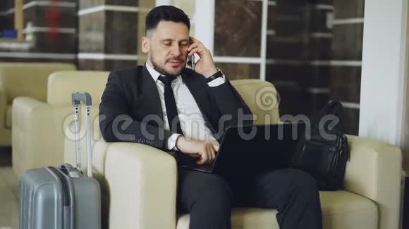 坐在豪华酒店的扶手椅上时自信的商人会用手机和笔记本电脑说话视频的预览图