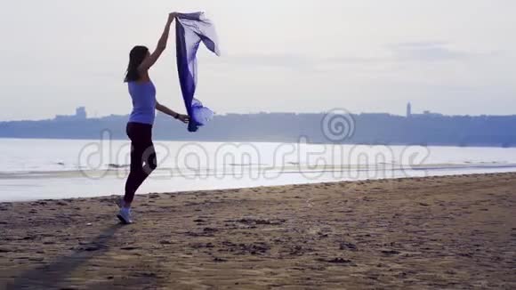 穿着运动服带着蓝色围巾跳舞的年轻苗条黑发女人一个女人在一个大丽的沙滩上订婚视频的预览图