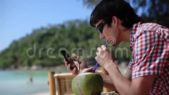 年轻的旅行者戴着太阳镜在海滩咖啡馆喝着新鲜的椰子汁背景和背景的棕榈树上有海景视频的预览图