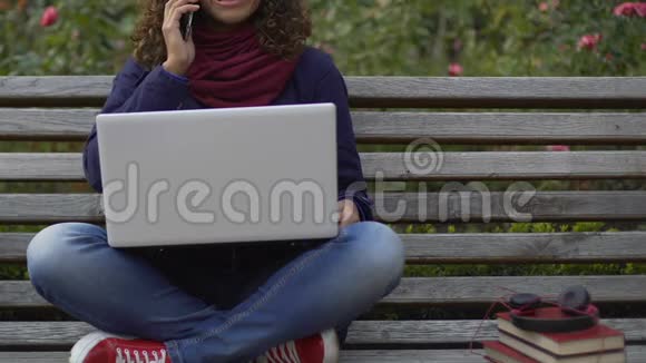 快乐的混血女孩坐在长凳上拿着笔记本电脑在智能手机上聊天视频的预览图