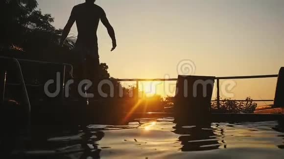 年轻人的剪影在夕阳下的游泳池里跳着穿过美丽的日落慢动作视频的预览图