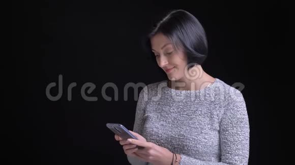 成人美丽的白种人女子的特写肖像在电话上浏览在黑色背景下笑视频的预览图