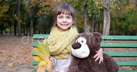 小女孩坐在长凳上手里拿着泰迪熊和树叶摆着背景秋公园视频的预览图