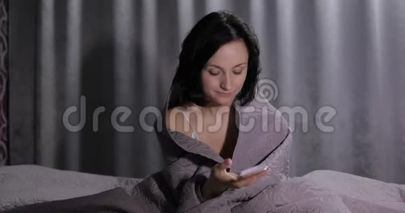 女人坐在毯子下面的床上享受着与智能手机上的朋友聊天视频的预览图