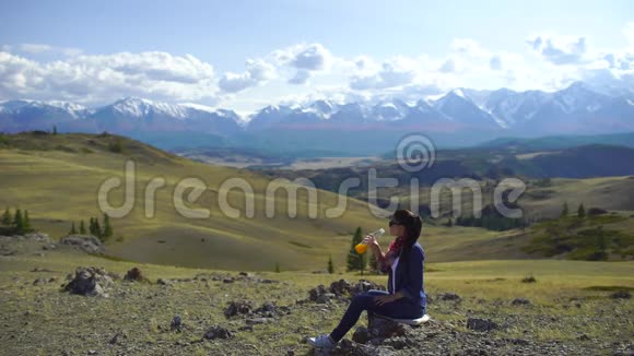年轻美丽的女旅行者在山顶上喝橙汁周围有山和小山4视频的预览图