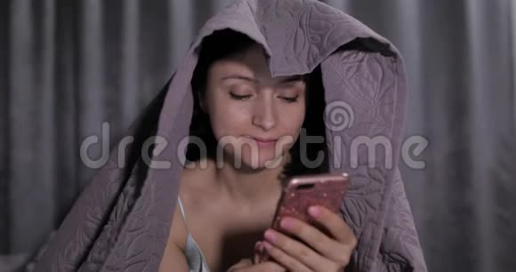 女人坐在毯子下面的床上享受着与智能手机上的朋友聊天视频的预览图