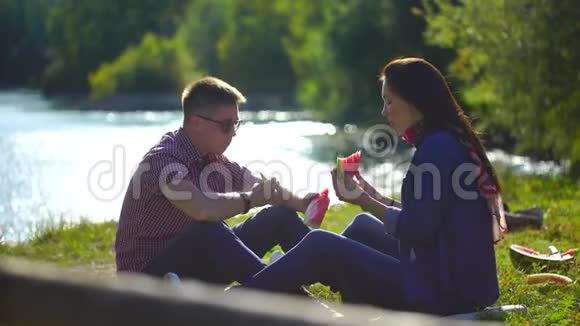 年轻人和女人在河边的森林里野餐吃西瓜背景是山4千克视频的预览图