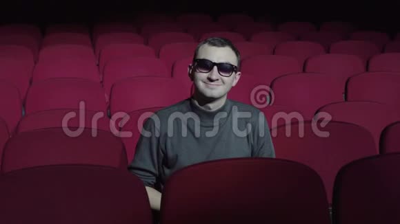 一个戴3D眼镜的单身男子坐在黑色电影院舒适的红色椅子上看着摄像机微笑着视频的预览图